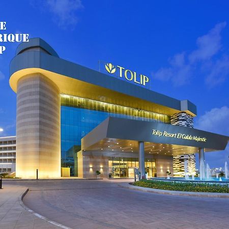 Tolip Resort El Galala Majestic Ain Sukhna Extérieur photo The photo shows a modern hotel building named "Tolip Resort & Spa," featuring a sleek architectural design with large glass windows. The hotel is illuminated against a twilight sky, showcasing its prominent signage. In the foreground, there is a well