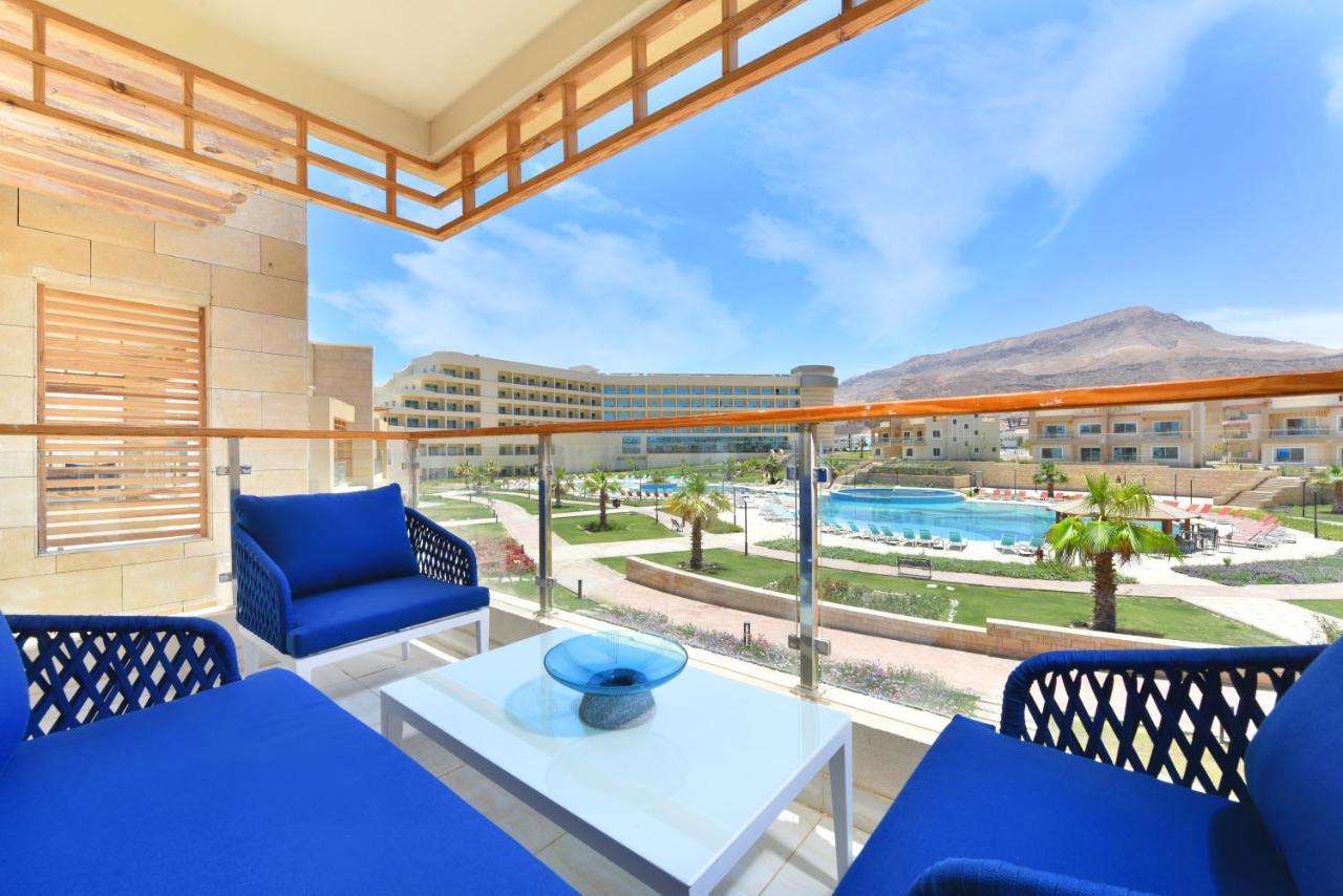 Tolip Resort El Galala Majestic Ain Sukhna Extérieur photo The photo showcases a balcony view featuring two blue chairs and a table with a decorative bowl. The balcony overlooks a landscape that includes a swimming pool and manicured lawns. In the background, there are multiple buildings and palm trees, all 