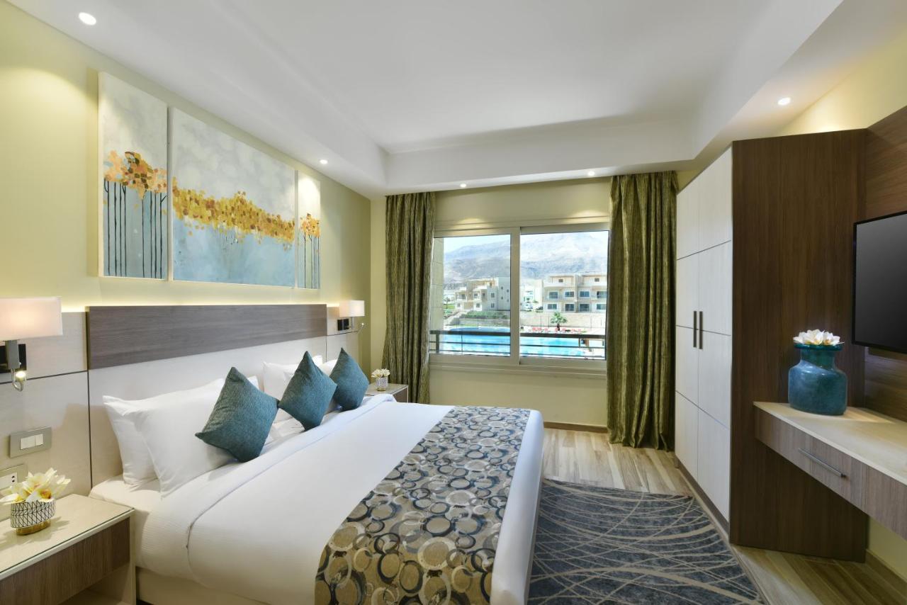 Tolip Resort El Galala Majestic Ain Sukhna Extérieur photo The photo shows a modern hotel room. It features a large bed with white bedding and several decorative pillows in shades of teal. Above the bed, there are two pieces of wall art with a landscape theme. The room has a large window with green curtains,