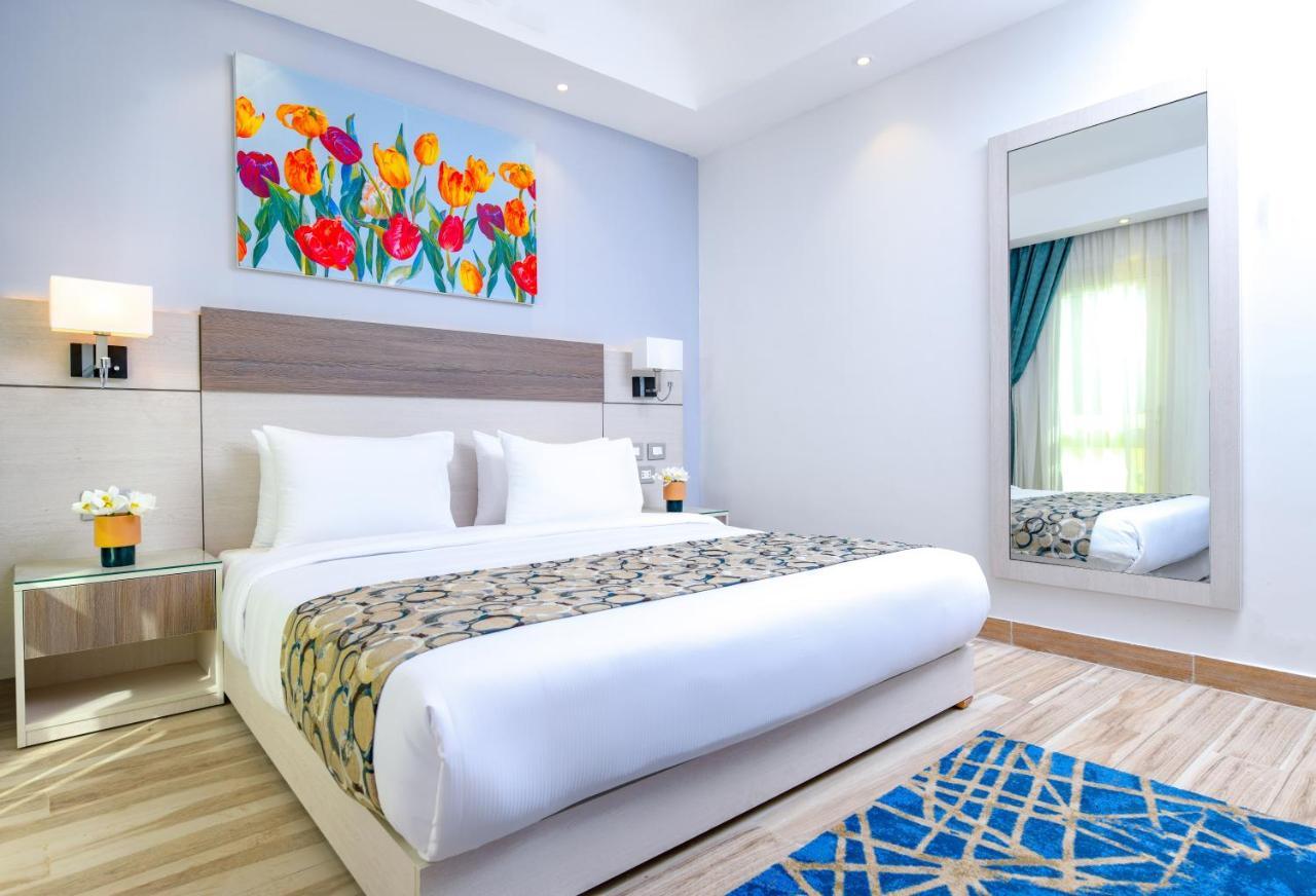Tolip Resort El Galala Majestic Ain Sukhna Extérieur photo The photo shows a modern hotel bedroom. It features a large bed with white linens and a decorative throw blanket. The room has light wooden flooring and is bright, with ample natural light coming from a window. There is a colorful painting of flowers