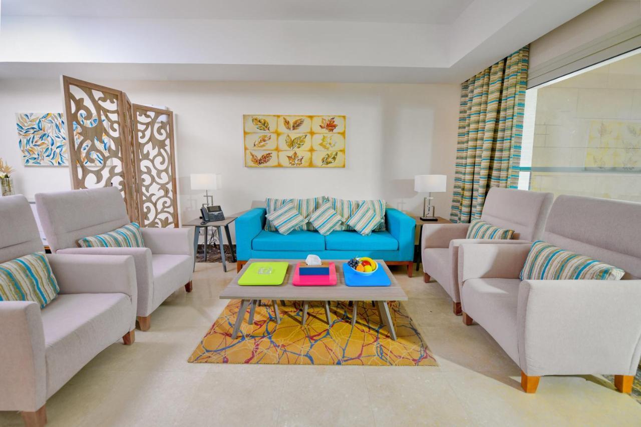 Tolip Resort El Galala Majestic Ain Sukhna Extérieur photo The photo shows a stylish and colorful living room. It features a bright blue sofa with patterned cushions, complemented by two light gray armchairs. In front of the sofa, there's a wooden coffee table with vibrant trays in pink, green, and yellow. A