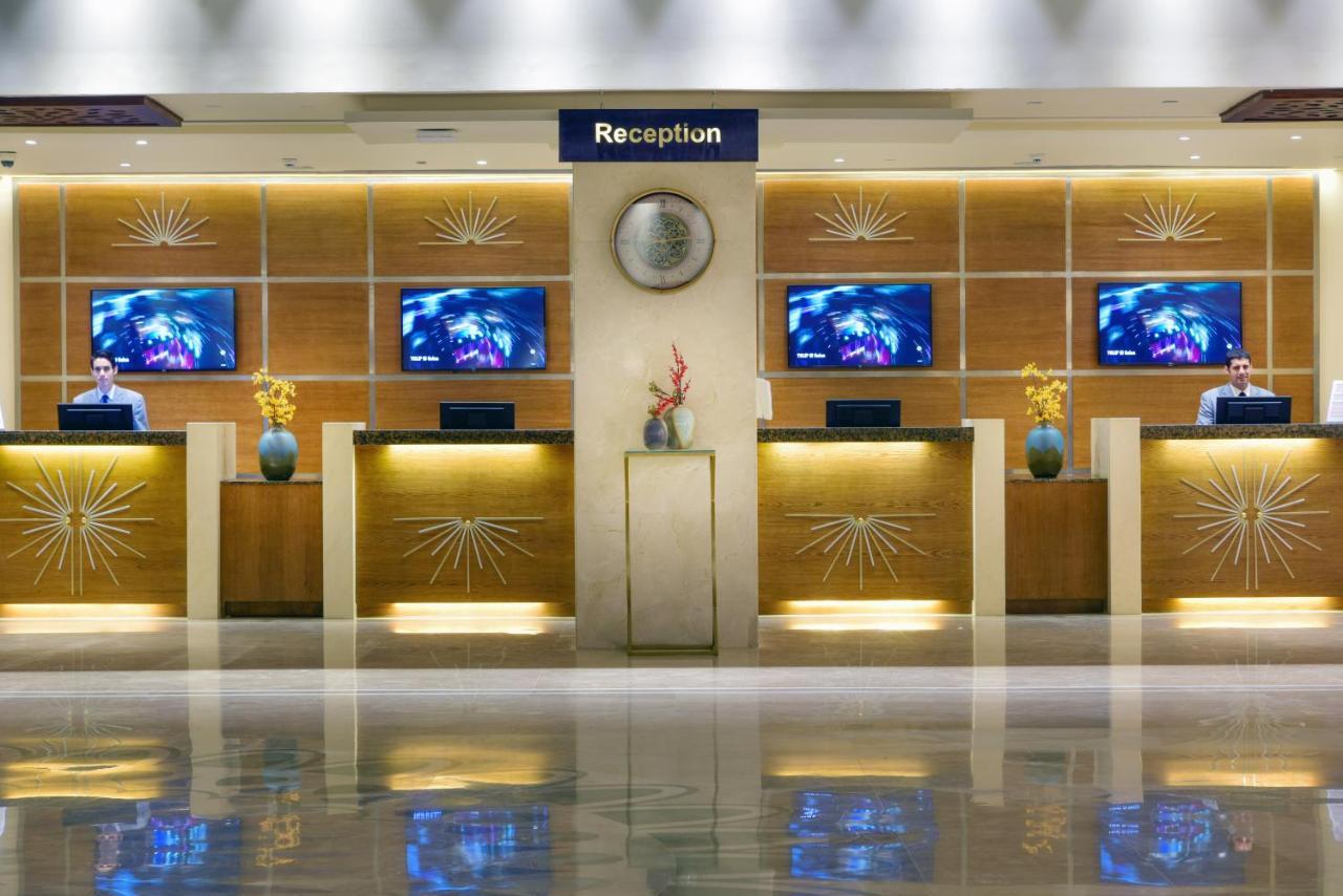 Tolip Resort El Galala Majestic Ain Sukhna Extérieur photo The image shows a hotel reception area. There are several reception desks lined up against a wall with decorative paneling. Each desk has a staff member, and above the desks, there are screens displaying information. A clock is visible on the wall, a