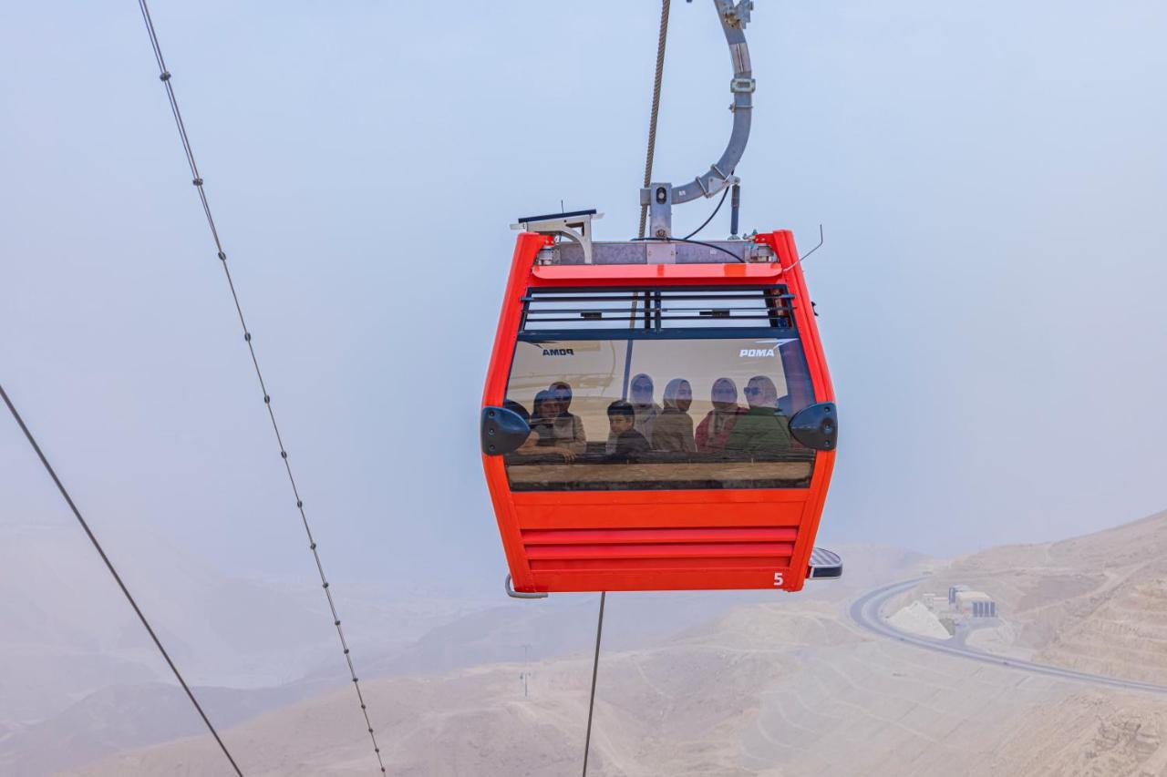 Tolip Resort El Galala Majestic Ain Sukhna Extérieur photo The photo shows a red gondola lift suspended in the air, traveling along cables. Inside the gondola, a group of people can be seen. The background features a hazy landscape, possibly mountains or hills, indicating an elevated area. The scene conveys 