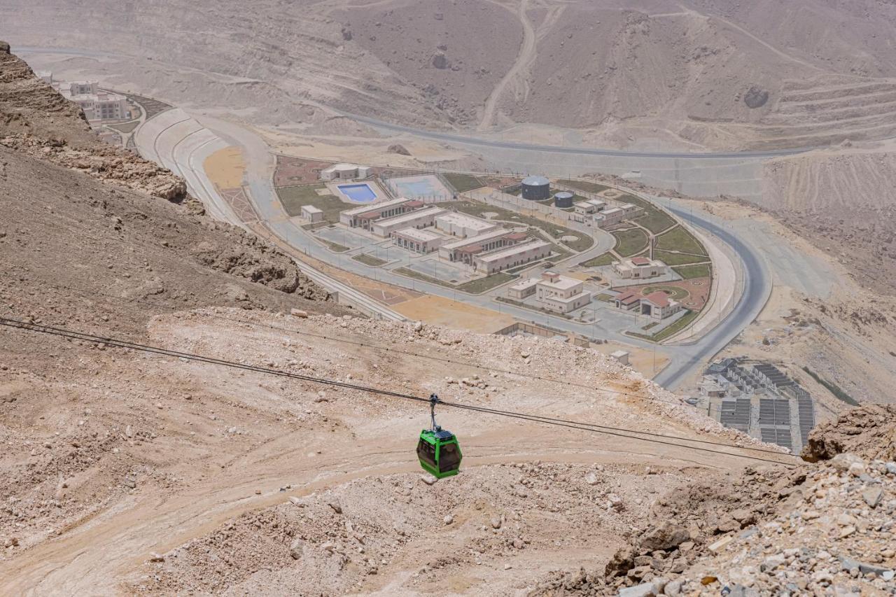 Tolip Resort El Galala Majestic Ain Sukhna Extérieur photo The photo shows a mountainous landscape with a steep, dry terrain. In the foreground, there is a green cable car or gondola traveling along a cable. Below, a complex of buildings and roads can be seen, likely part of a resort or recreational area. Th
