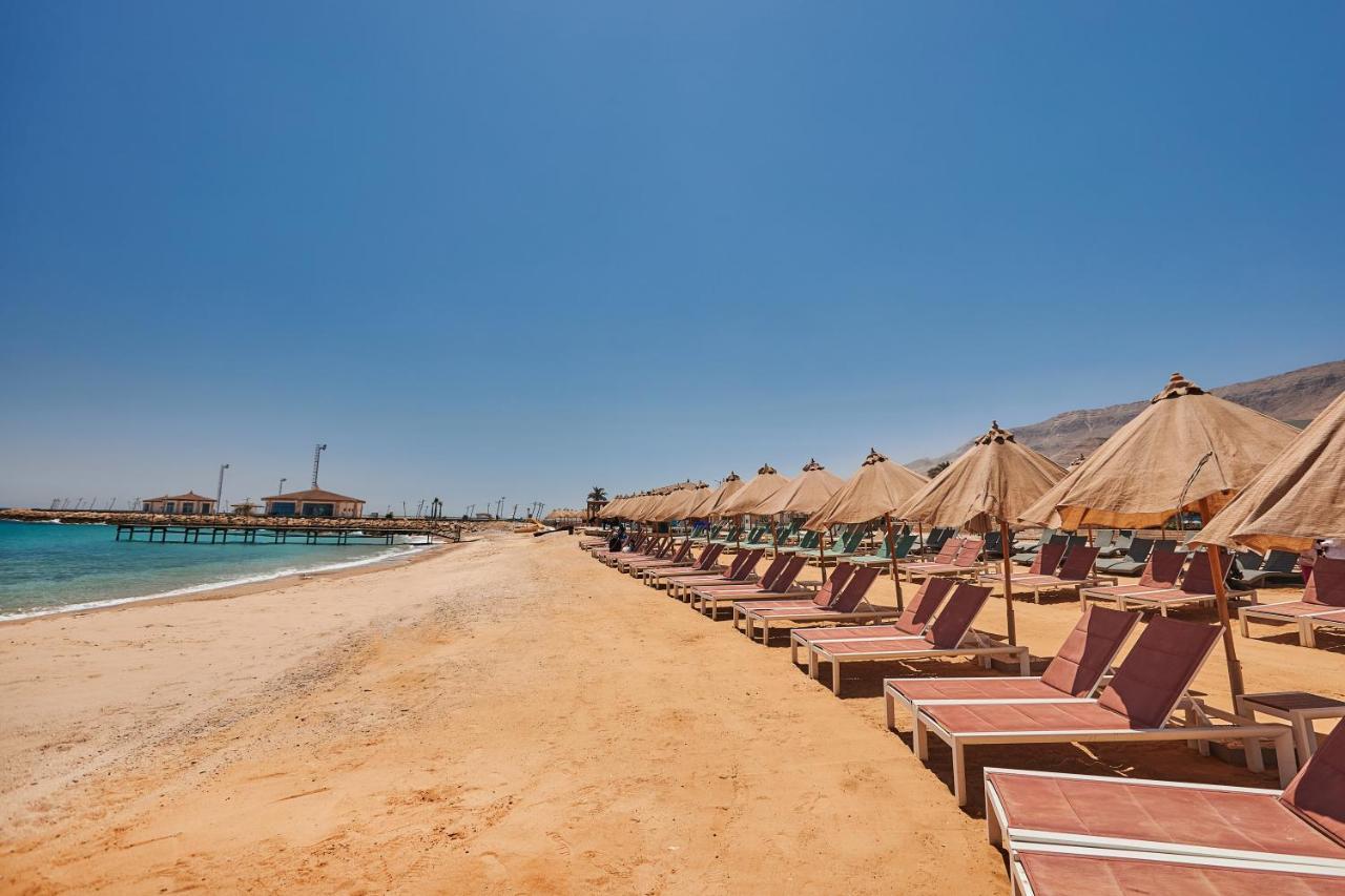 Tolip Resort El Galala Majestic Ain Sukhna Extérieur photo The photo shows a scenic beach with a stretch of golden sand under a clear blue sky. There are several beach loungers arranged in rows, each accompanied by thatched umbrellas providing shade. In the background, there is a wooden pier extending into t