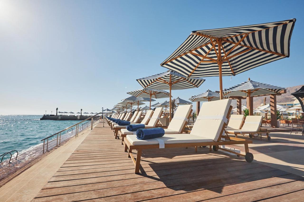Tolip Resort El Galala Majestic Ain Sukhna Extérieur photo The photo depicts a serene beachside setting featuring a row of sun loungers arranged on a wooden deck. Each lounger is equipped with a folded towel and shaded by striped umbrellas. In the background, the water sparkles under bright sunlight, and the