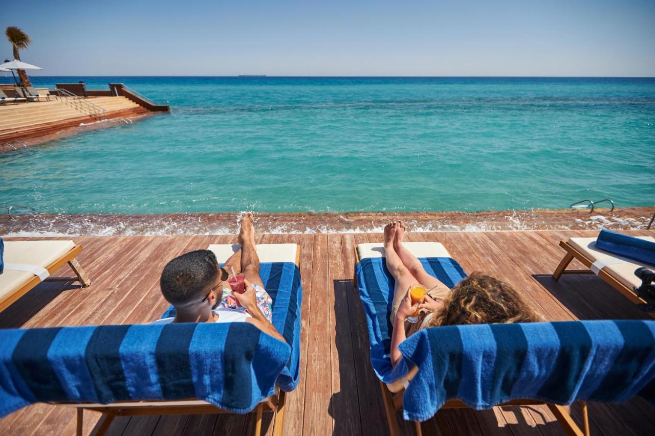 Tolip Resort El Galala Majestic Ain Sukhna Extérieur photo The photo depicts two people relaxing on lounge chairs by the waterfront. They are positioned on a wooden deck with a view of the clear, turquoise water. Both individuals are reclining, enjoying the sun, and each has a drink in hand. The scene convey