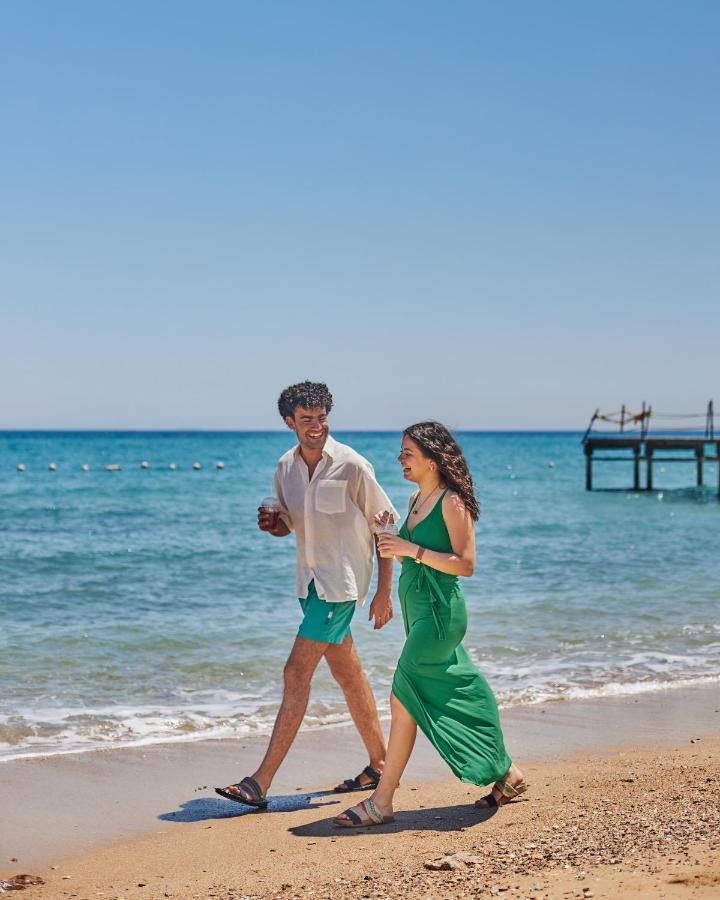 Tolip Resort El Galala Majestic Ain Sukhna Extérieur photo The photo shows a couple walking along a beach. The man is wearing a white short-sleeved shirt and green shorts, while the woman is dressed in a flowing green dress. They both appear to be enjoying the pleasant weather, with the ocean in the backgrou
