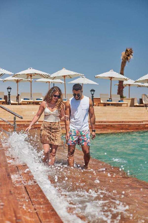 Tolip Resort El Galala Majestic Ain Sukhna Extérieur photo The photo depicts a couple walking along a poolside area. They are enjoying a sunny day near a swimming pool, with splashes of water around them. The woman is wearing a light-colored top and a patterned skirt, while the man is dressed in a sleeveless