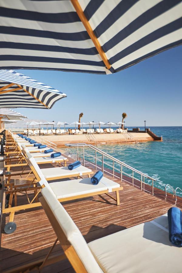 Tolip Resort El Galala Majestic Ain Sukhna Extérieur photo The photo depicts a serene outdoor area by the waterfront. There are several lounge chairs arranged in a row, each equipped with blue towels. Large, striped umbrellas provide shade over some of the chairs. In the background, there is a wooden deck th