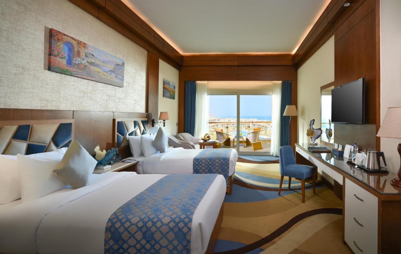 Tolip Resort El Galala Majestic Ain Sukhna Extérieur photo The photo shows a well-decorated hotel room featuring two beds with decorative pillows. The room has wooden accents and modern furnishings, including a desk, a television, and lamps. Large windows flood the space with natural light, offering a view o