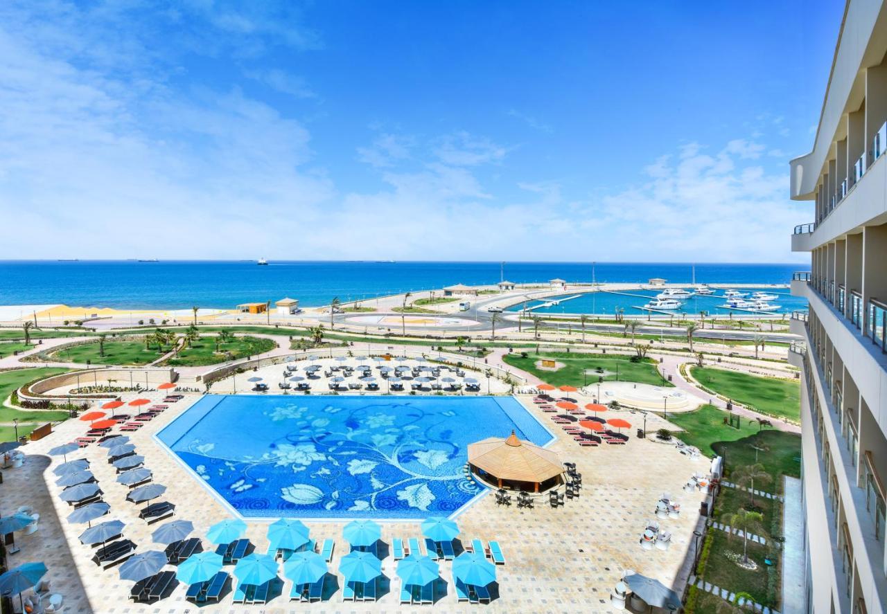 Tolip Resort El Galala Majestic Ain Sukhna Extérieur photo The photo shows a beautiful outdoor view of a resort area featuring a large swimming pool with a decorative underwater design. Surrounding the pool are numerous sun loungers and umbrellas in a variety of colors. In the background, there is a scenic c
