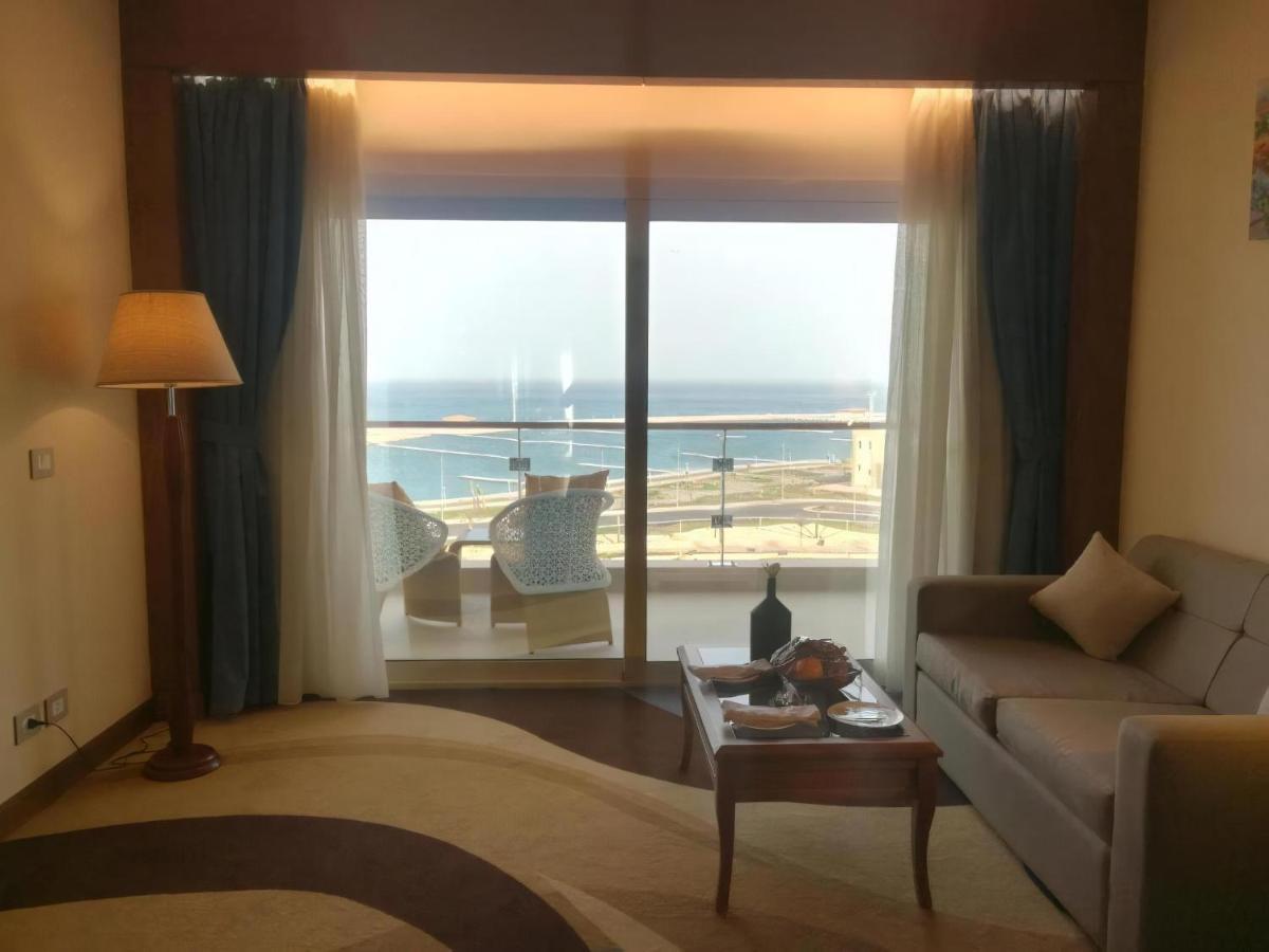 Tolip Resort El Galala Majestic Ain Sukhna Extérieur photo The photo shows a stylish interior of a room with a large window overlooking the sea. The window features sheer and thick curtains and is framed by a balcony with two chairs visible outside. Inside, there is a comfortable-looking sofa, a coffee table
