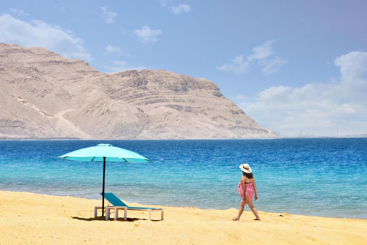 Tolip Resort El Galala Majestic Ain Sukhna Extérieur photo The photo depicts a serene beach scene. In the foreground, there is a sandy shore with a single beach chair and an umbrella in a light blue color. A person dressed in a pink outfit is walking along the shoreline, enjoying the beach atmosphere. In the